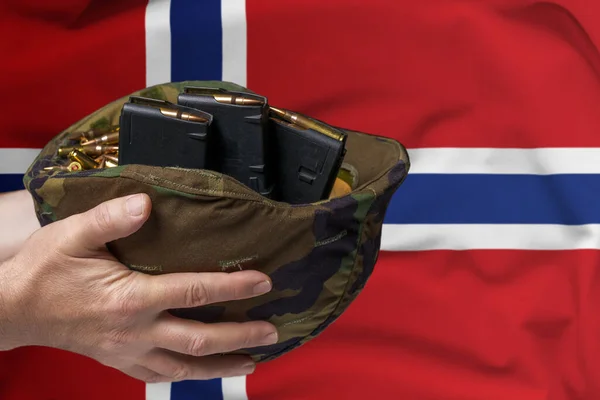 A military helmet with cartridges and magazines for a rifle in the hands of a man against the background of the flag of Norway. The concept of selling weapons or military assistance.