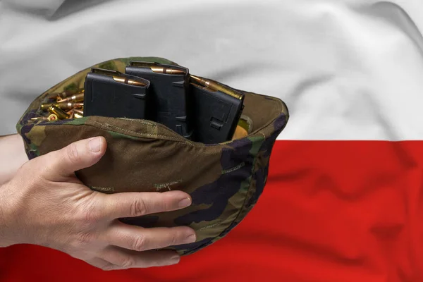 A military helmet with cartridges and magazines for a rifle in the hands of a man against the background of the flag of Poland. The concept of selling weapons or military assistance.