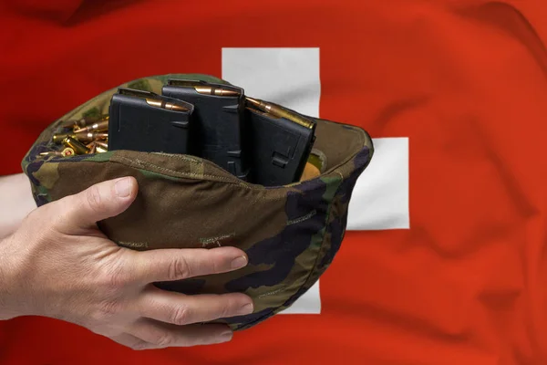A military helmet with cartridges and magazines for a rifle in the hands of a man against the background of the flag of Switzerland. The concept of selling weapons or military assistance.