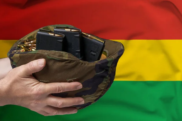 A military helmet with cartridges and magazines for a rifle in the hands of a man against the background of the flag of Bolivia. The concept of selling weapons or military assistance.