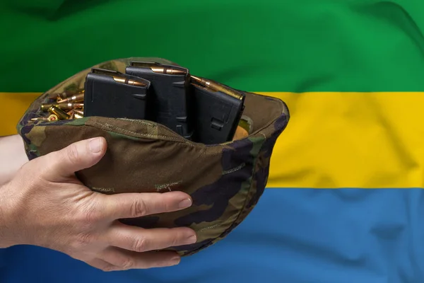 A military helmet with cartridges and magazines for a rifle in the hands of a man against the background of the flag of Gabon. The concept of selling weapons or military assistance.