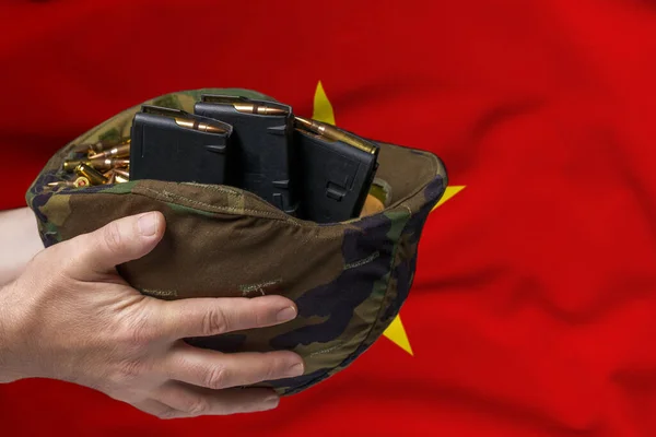 A military helmet with cartridges and magazines for a rifle in the hands of a man against the background of the flag of Vietnam. The concept of selling weapons or military assistance.
