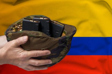 Fişek ve şarjör dolu bir askeri miğfer. Kolombiya bayrağının arkasına dayanmış bir adamın elinde tüfek. Silah ya da askeri yardım satma kavramı.