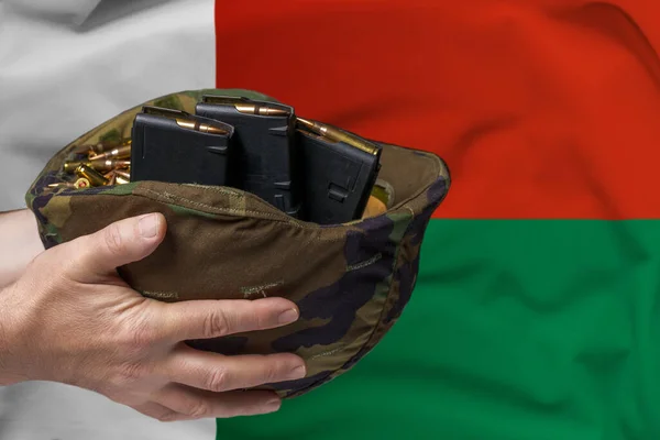 A military helmet with cartridges and magazines for a rifle in the hands of a man against the background of the flag of Madagascar. The concept of selling weapons or military assistance.