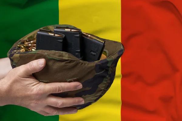 A military helmet with cartridges and magazines for a rifle in the hands of a man against the background of the flag of Mali. The concept of selling weapons or military assistance.