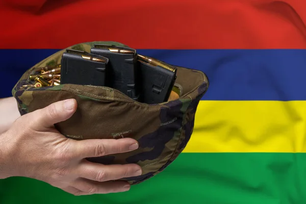 A military helmet with cartridges and magazines for a rifle in the hands of a man against the background of the flag of Mauritius. The concept of selling weapons or military assistance.