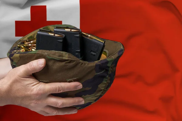 A military helmet with cartridges and magazines for a rifle in the hands of a man against the background of the flag of Tonga. The concept of selling weapons or military assistance.