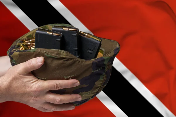 A military helmet with cartridges and magazines for a rifle in the hands of a man against the background of the flag of Trinidad and Tobago. The concept of selling weapons or military assistance.