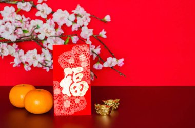Red envelope packet or ang bao (word mean wealth) puts with oranges and ingots with peach blossom on red glitter paper background. 