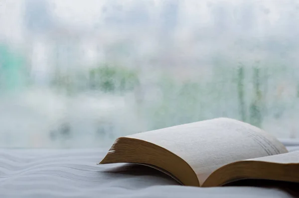 本は窓に雨が降って朝にベッドに置かれる 家にいてリラックスしたコンセプト ストック画像