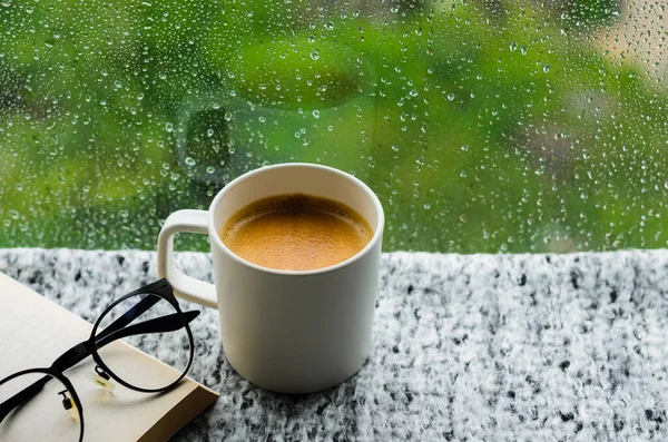 A cup of hot coffee with book and spectacles on table in morning with rain drop on window and green background. Stay home and relaxing concept.