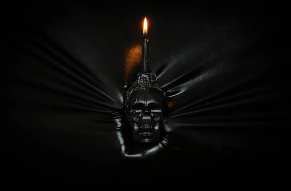 Black candle with flame on skull holding by hand pressing through black fabric background. Halloween festival concept.