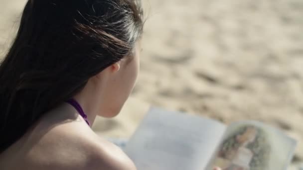Vista posterior de la joven leyendo libro sobre la playa de arena — Vídeos de Stock