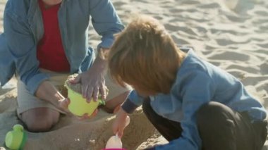 Çocuk kumsalda kürekle kovada kum topluyor.