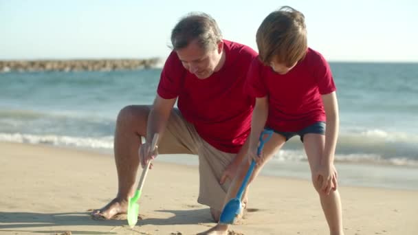 Largo tiro de padre soltero dibujo en la arena con hijo en la playa — Vídeo de stock