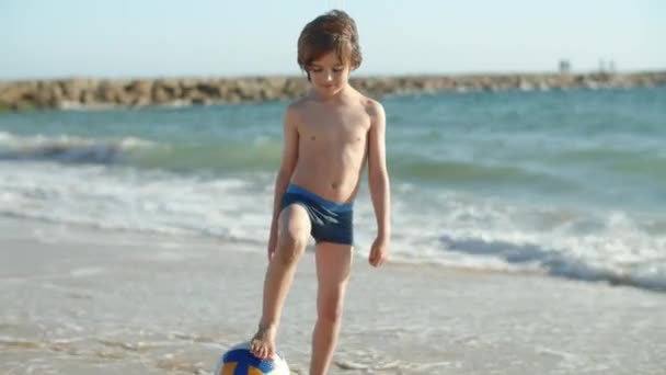 Elölnézetben a fiú áll a homokos tengerparton lábával a labdát — Stock videók