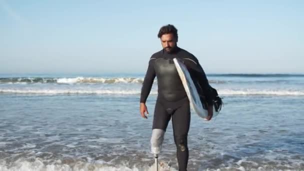 Vista frontal de surfista serio con pierna biónica saliendo del océano — Vídeos de Stock