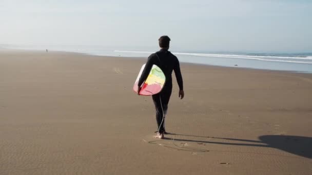 Colpo lungo di surfista con gamba artificiale che cammina lungo la spiaggia — Video Stock