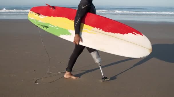 Человек с искусственной ногой идет вдоль пляжа с доской для серфинга — стоковое видео