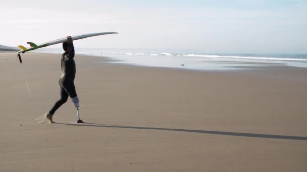 Dalekie ujęcie niepełnosprawnego człowieka idącego do oceanu z deską surfingową — Wideo stockowe