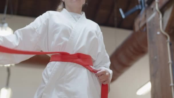 Mediana toma de chica atando cinturón rojo en kimono en sala de práctica — Vídeos de Stock