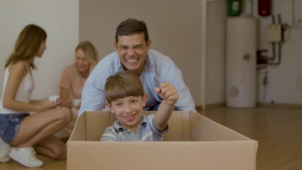 Padre empujando hijo sentado en caja alrededor de la casa después de mover — Vídeo de stock