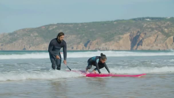 Largo tiro de chica feliz de pie en la tabla de surf con la ayuda del entrenador — Vídeos de Stock