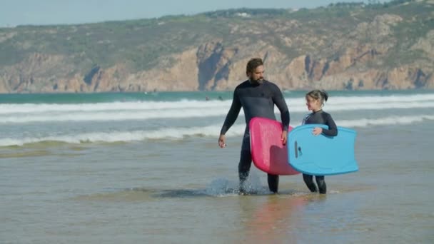 Вид спереди тренера по серфингу с инвалидностью и ребенком, заходящим в океан — стоковое видео
