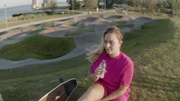 Vacker flicka sitter på gräsmattan i skate park och dricksvatten — Stockvideo