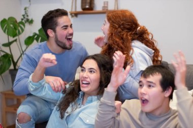 Televizyon seyreden heyecanlı arkadaşlar, futbol takımının galibiyetini kutluyorlar. Tutkulu sporseverler oturma odasında koçun üzerinde oturuyorlar. Mutlu erkekler ve kadınlar çığlık atıyor, futbol takımı için tezahürat yapıyorlar. Zafer, duygu konsepti