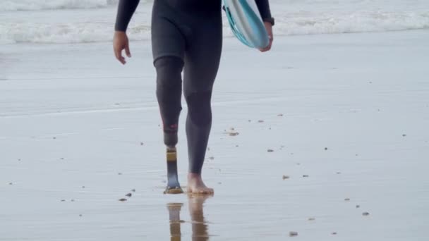 Hombre con pierna artificial caminando por la orilla mojada con tabla de surf — Vídeos de Stock
