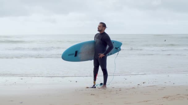 Hombre con pierna biónica de pie en la playa con tabla de surf bajo el brazo — Vídeos de Stock