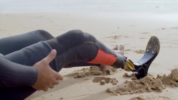 Surfista con discapacidad sentado en la orilla y amasando las piernas — Vídeos de Stock