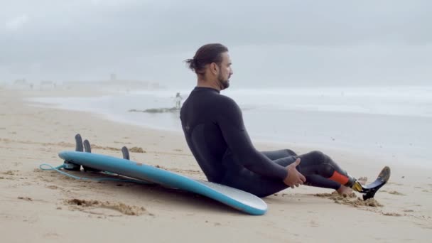 Surfista cansado con pierna biónica sentado en la orilla del océano — Vídeos de Stock