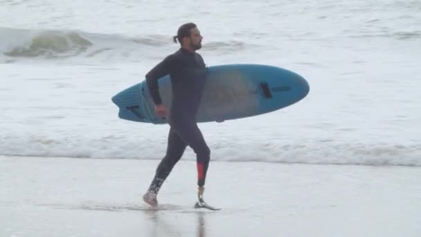 Handsome man running along coast with paddle board under arm — Stock Video