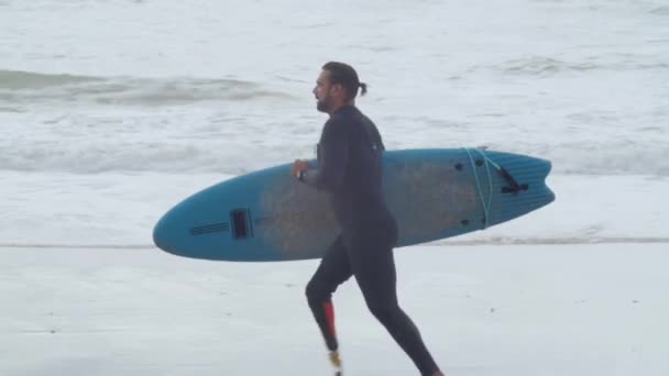 Hombre con pierna artificial corriendo por la costa con tabla de surf — Vídeos de Stock