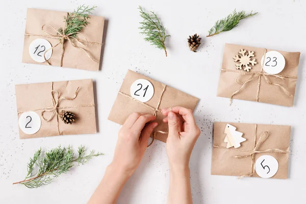 Calendário do advento do Natal. Mãos de mulheres que fazem presentes com envelopes de kraft, ramos de abeto, cones de pinheiro. Vista superior Imagens De Bancos De Imagens