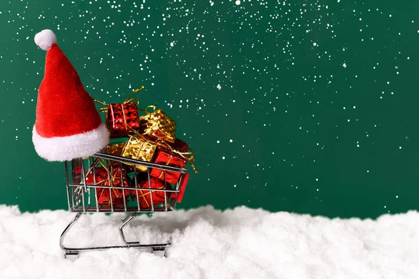 Carrito de compras, sombrero de santa, regalos sobre hermoso fondo verde con nieve. Vista frontal. Navidad, concepto de año nuevo. Copiar espacio —  Fotos de Stock