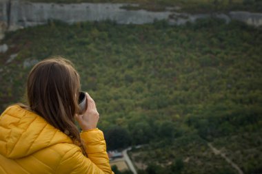Genç bayan gezgin çay dağı. Güzel bir sonbahar ormanıyla bir dağın tepesinde oturan bir kız. Seyahat etme, doğanın tadını çıkarma, mahremiyet, rahatlama. Sıcak içecek elleri yakın.