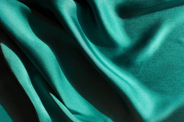 Mavi-yeşil saten kumaş. Masanın üzerinde düz ipek kumaş kesilmiş. Seçici odak noktasında parlak yumuşak kıvrımlar. Deniz dalgasının koyu turkuaz rengi. Kapaklar, web siteleri, dikiş atölyeleri için zengin tasarım.