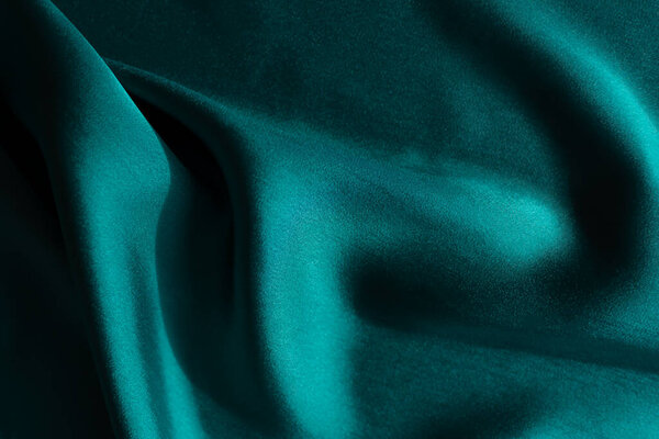 Blue-green satin fabric. Smooth silk fabric cut off on the table. Shiny soft folds in selective focus. Dark turquoise color of the sea wave. Deep rich design for covers, websites, sewing workshops.