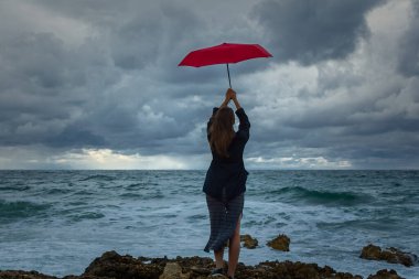 Denizin arka planında kırmızı şemsiyeli genç bir kadın. Atmosferik sonbahar manzarası gelişmekte olan elbiseli, koyu renk saçlı genç bir kızla. Fırtına, güçlü rüzgar, bulutlu. Mutluluk kavramı