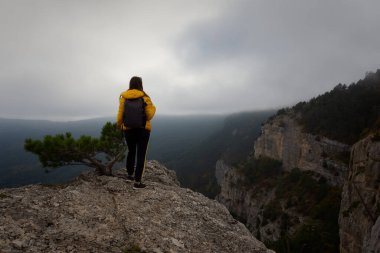 Young dark-haired woman backs mountains autumn landscape. Cloudy gray foggy view down the mountain. A happy tourist with a backpack. The concept of an active lifestyle, ecotourism. Alone with nature