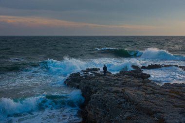Deniz fırtınası plajı. Element büyük dalgalar taştan kıyıda kırılır. Yaklaşan bir fırtınayla deniz manzarası. Bir adam öfkeli dalgaların arka planında poz veriyor. Tatil köyü kavramı, iklim değişiklikleri