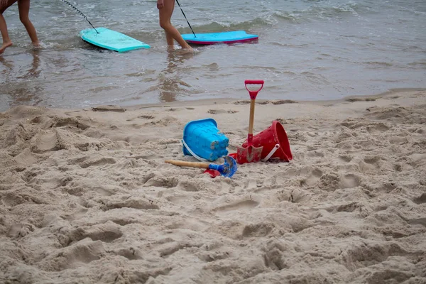 colorful plastic beach shovels and pails on the seashore with kids dragging boogie boards in the water