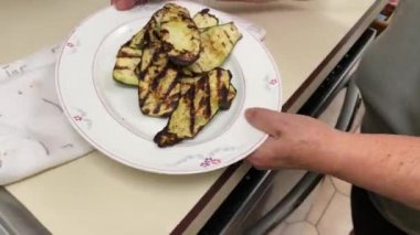 Bir kadın ızgarada taze ızgara kabak pişiriyor ve gümüş bir spatulayla birlikte beyaz servis tabağına yerleştiriliyor.
