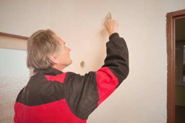 Older man removing old wallpapers off the walls in empty apartment