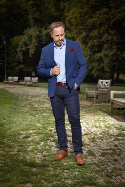 Mavi takım elbiseli ve kravatlı bir adam yaz parkında poz veriyor.