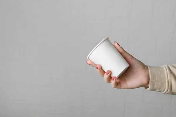 Mano femminile che tiene vuoto vuoto portare via tazza di carta su sfondo neutro parete grigia — Foto Stock