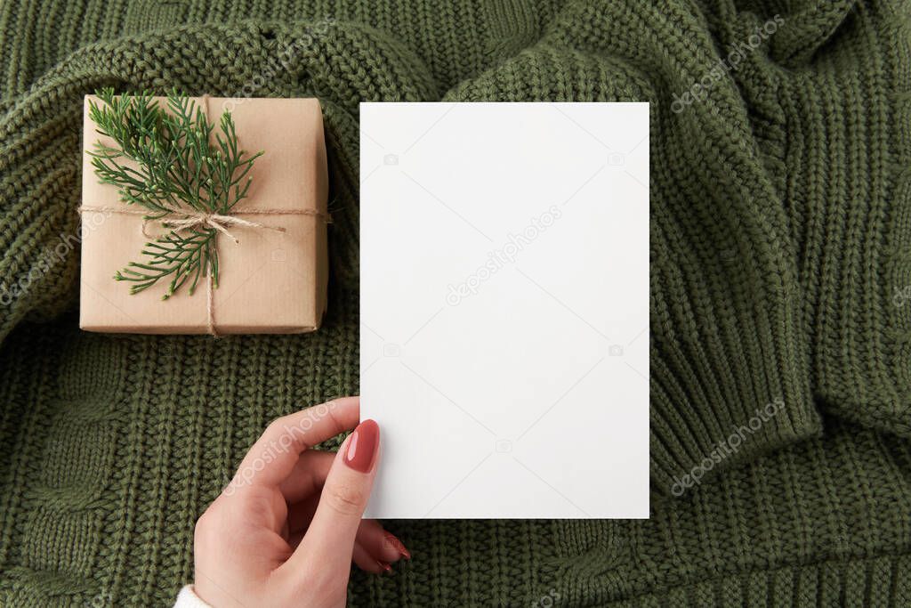 empty 5x7 card mockup kraft gift box with fir decoration on knitted green sweater, design element for wedding invitation, thank you or greeting card. Christmas winter card mock up background